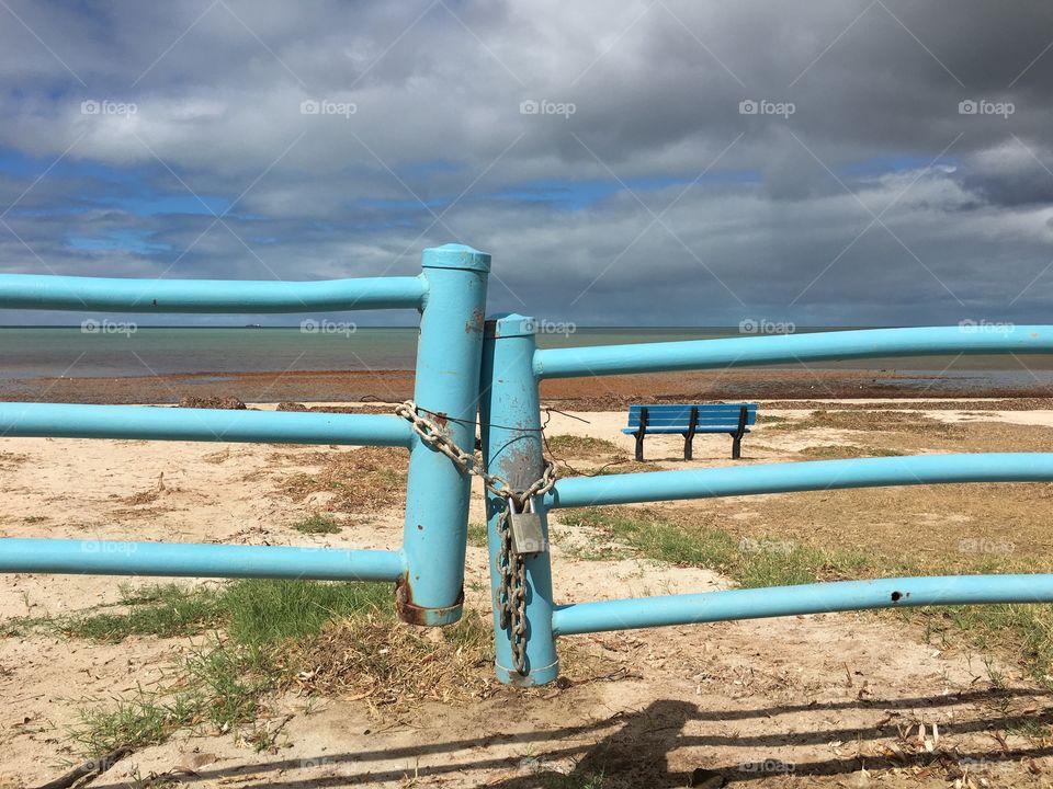 Turquoise aqua blue metal gate barrier to beach, with chain and padlock on stormy day and blue bench in background 