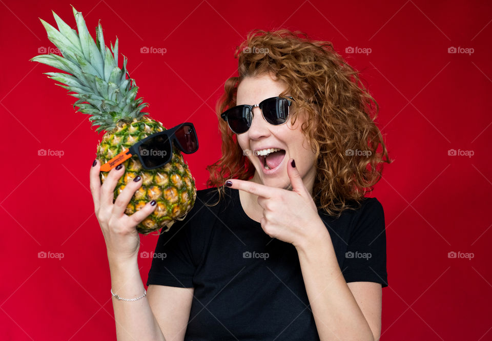 beautiful red-haired girl, in a black T-shirt with a pineapple in her hands, on a red background, has fun and frolics. Summer mood. Sexy happy girl