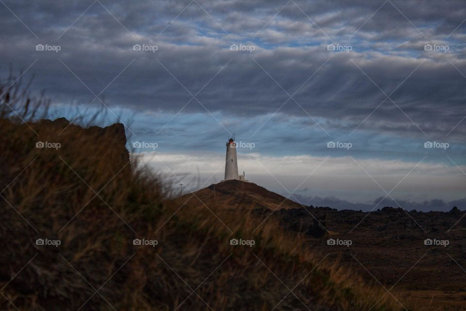 Lonely lighthouse in Iceland with hill and grass in the foreground and cloudy sky looking a bist stormy and loomy.