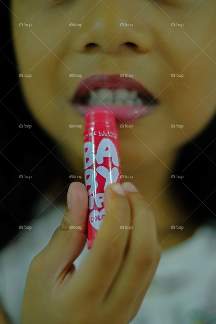 Cropped view of young girl holding lip balm near her lips isolated with dark background.