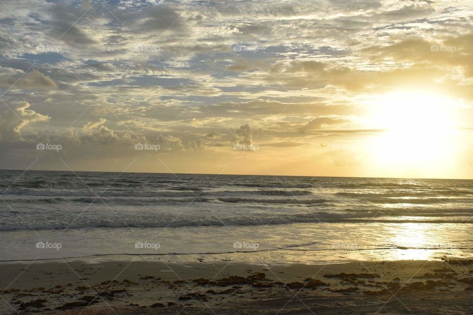 Stunning sunset over Daytona Beach in Florida. The bright, yellow sun rays stretch across the blue, cloudy sky and reflect on the ocean waves.