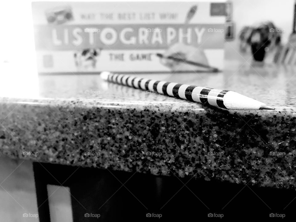 Fun black and white photo close up of striped pencil in forefront and Listography board game in background. 