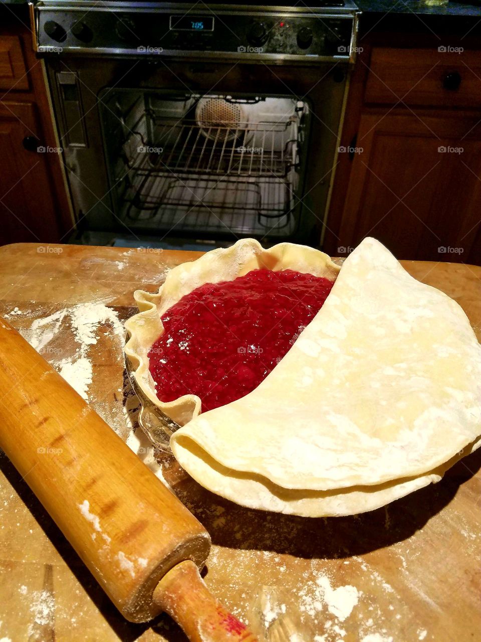 Raspberry Pie with top crust ready to lay flat, rolling pin, flour on board. Oven door open in background.