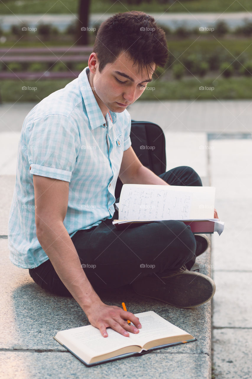 Student making the notes learning from books sitting on a monument outside of university. Young boy wearing a blue shirt and dark jeans