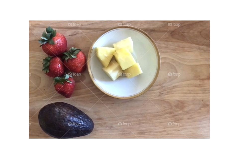 Three healthy fruits avocado, strawberries, and pineapples - green, red and yellow colorful fruits to eat for a healthy lifestyle - displayed on a wooden cutting board and plate