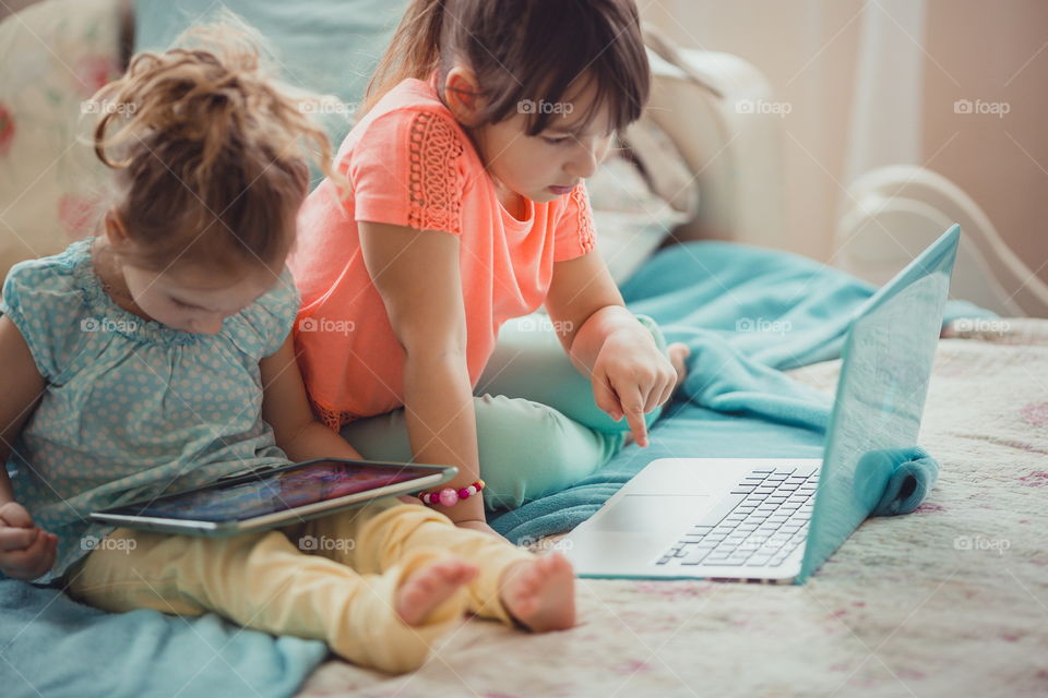 Little sisters with gadgets(laptop and tablet) in the bed.