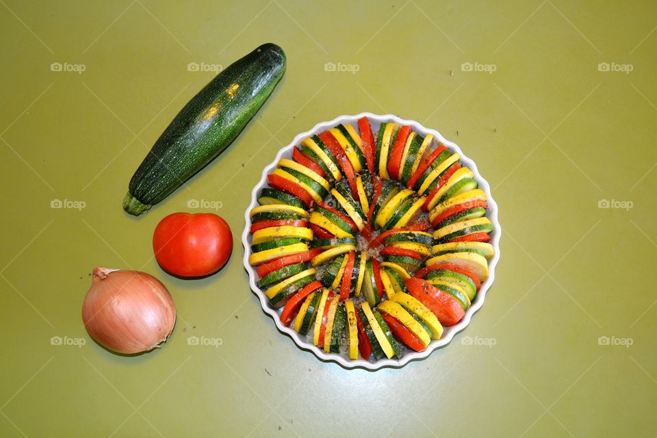 Vegetable Tian, A dish of fresh sliced zucchini, summer or yellow squash, potatoes and tomatoes arranged on a bed of diced onions. This Tian is arranged and ready to be covered with shredded cheese and baked.