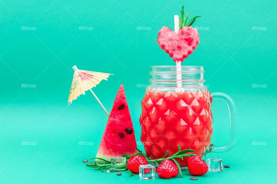 Watermelon-strawberry drink in a faceted jar with a handle, a metal lid, straws and fresh strawberries, pieces of watermelon in the shape of a heart and a triangle, ice cubes, roschmarine branches, bones and a skewer, a paper umbrella on a green back