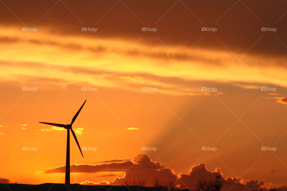 Impressive view of a wind turbine on the horizon at sunset. Orange-gold sky with some backlit clouds projecting their shadow upwards. Shot taken in Salento, Puglia, Italy.
