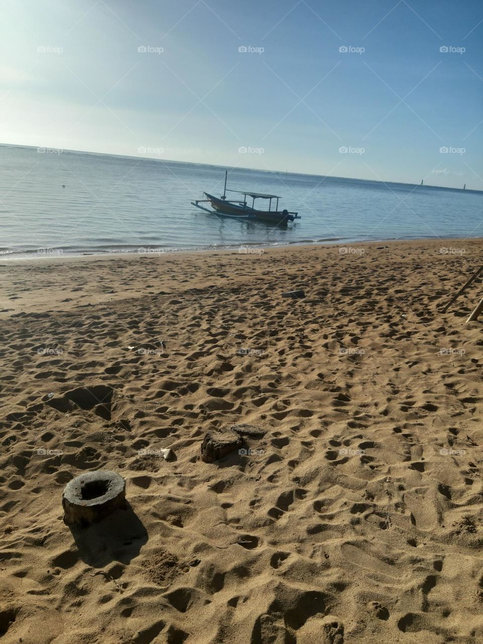 The beautiful morning view of sunrise seen from the brown soft sandy beach known as "Sanur Beach" which is located in Bali Island. In addition, there is a wooden fishing boat by the beach. It can tell us that most local people work as fisherman.