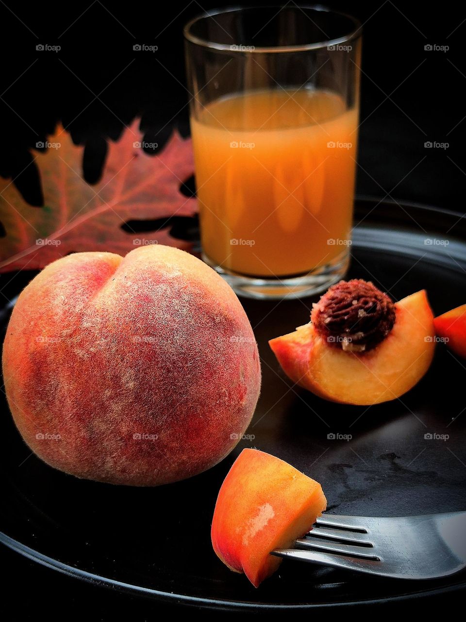 Fruit.  All shades of autumn in peach.  On a black plate lies a ripe peach, half a peach with a pit, and a glass of peach juice.  In the foreground is a fork with a piece of juicy peach.  In the background is an autumn oak leaf.  Black background