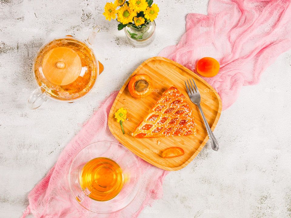 Beautiful view of one piece of apricot pie in a square wooden plate with a glass teapot and a mug of tea lie on a light cement background with a pink gauze napkin, flat lay close-up.