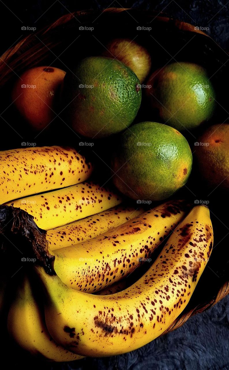 A rustic basket filled with ripe bananas and vibrant citrus fruits. The bananas, with their dark spots, signal sweetness and softness, while the oranges and green lemons contrast with bright hues. A play of light and shadow.