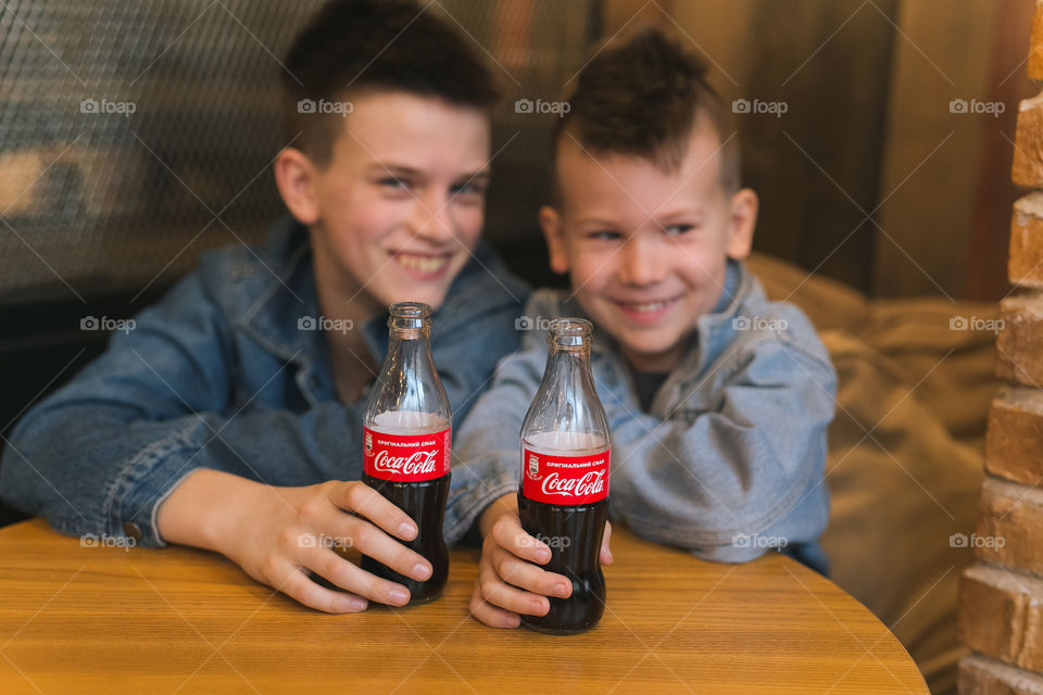 two lovely brothers, dressed in denim clothes, with beautiful hairstyles, are sitting in a cafe and drinking Coca-Cola.  communicate, laugh, smile.  two guys, two friends