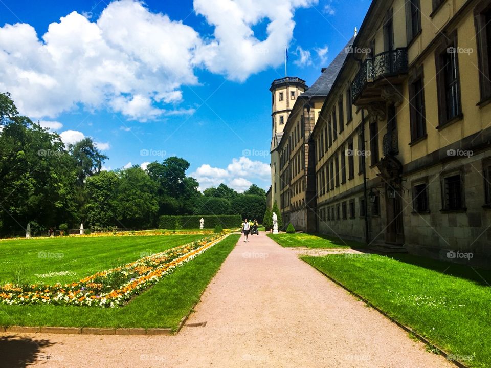 Castle in the city, with beautiful architecture, outside there are green grass and planted a variety of flowers. There are many people taking a walk, and also the couple are taking wedding photos