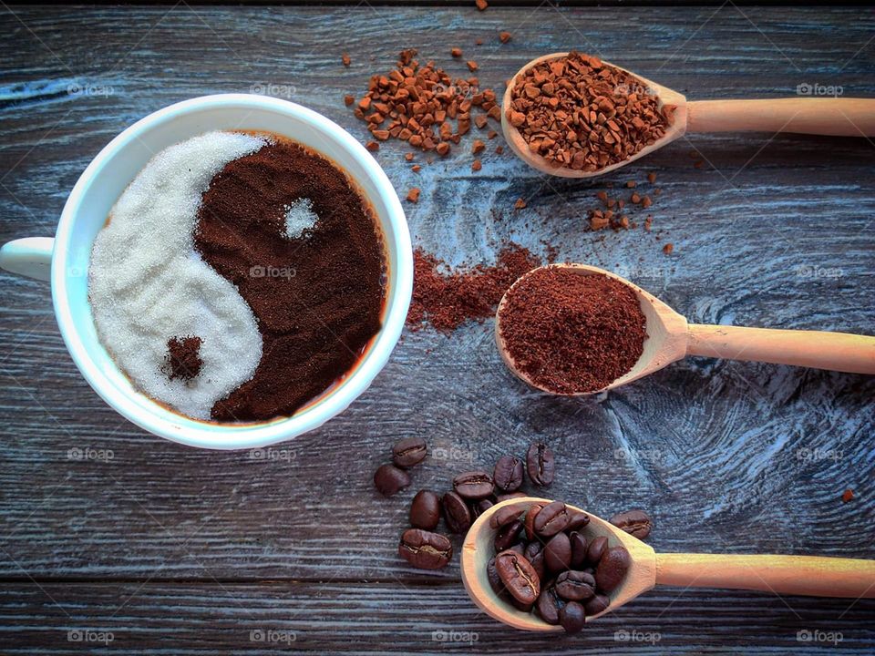 A white cup with hot black coffee with a yin-yang shape of white sugar on the surface and brown ground coffee. In front of the cup are three wooden spoons with different types of coffee: coffee beans, ground coffee and granulated coffee. The backgrou