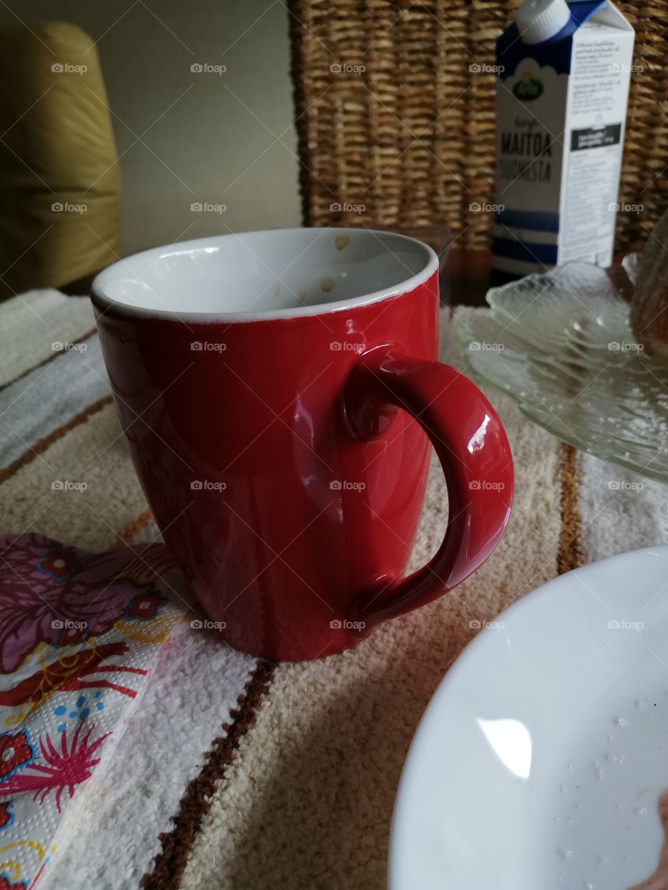 A red mug on a white, beige and brown striped table cloth. A clear decorated plate, a white one with some sugar, a patterned napkin and a milk carton on a brown wooden table. Couple of coffee spots inside the cup. Behind a back of a weaved chair