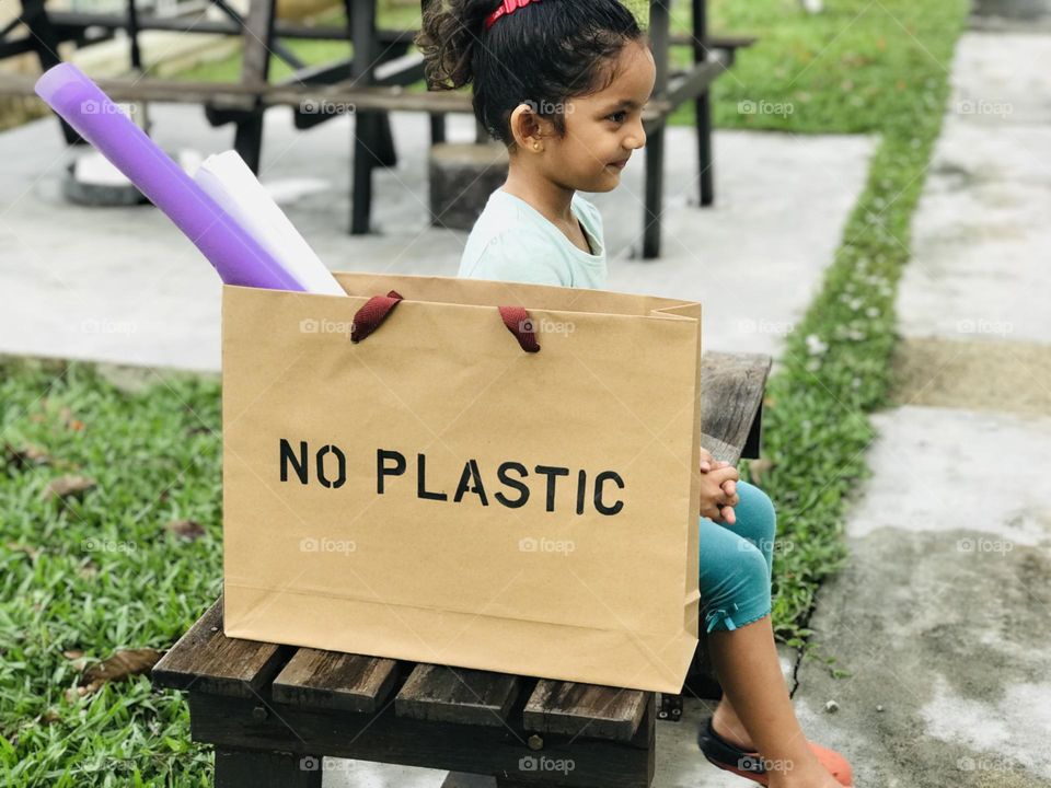 A baby girl sat on a bench and smile’s besides her a paper bag wrote as no plastic on its top.