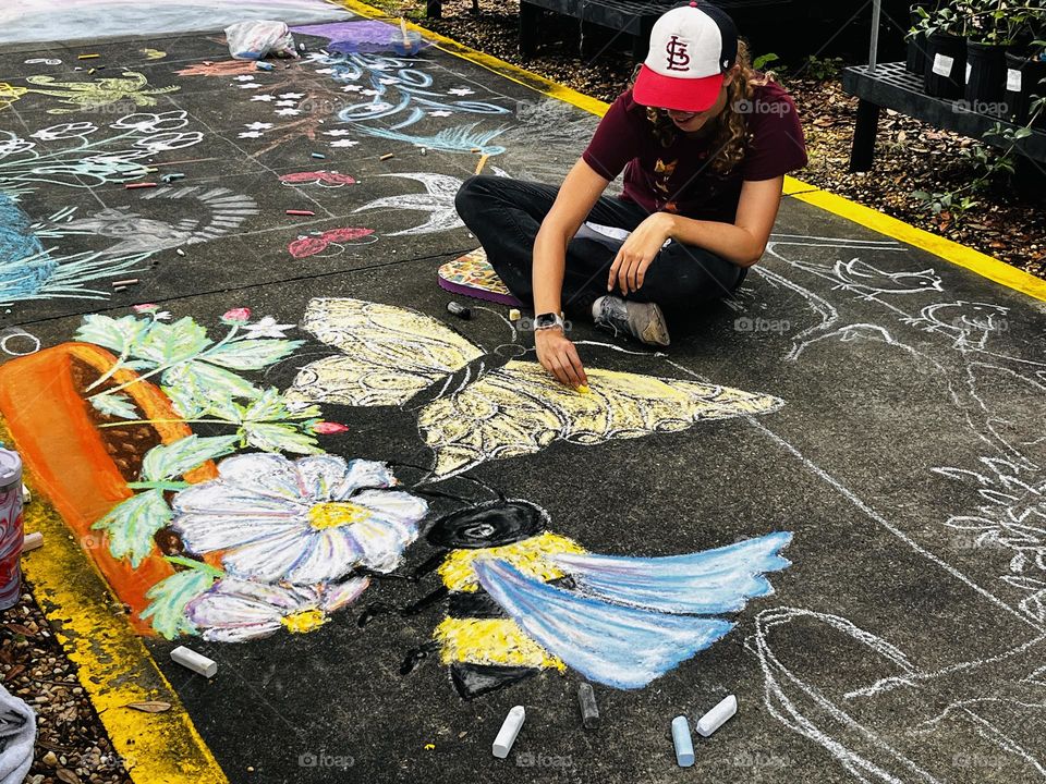Person using colored chalk to draw a floral scene on a sidewalk with a bee and butterfly 