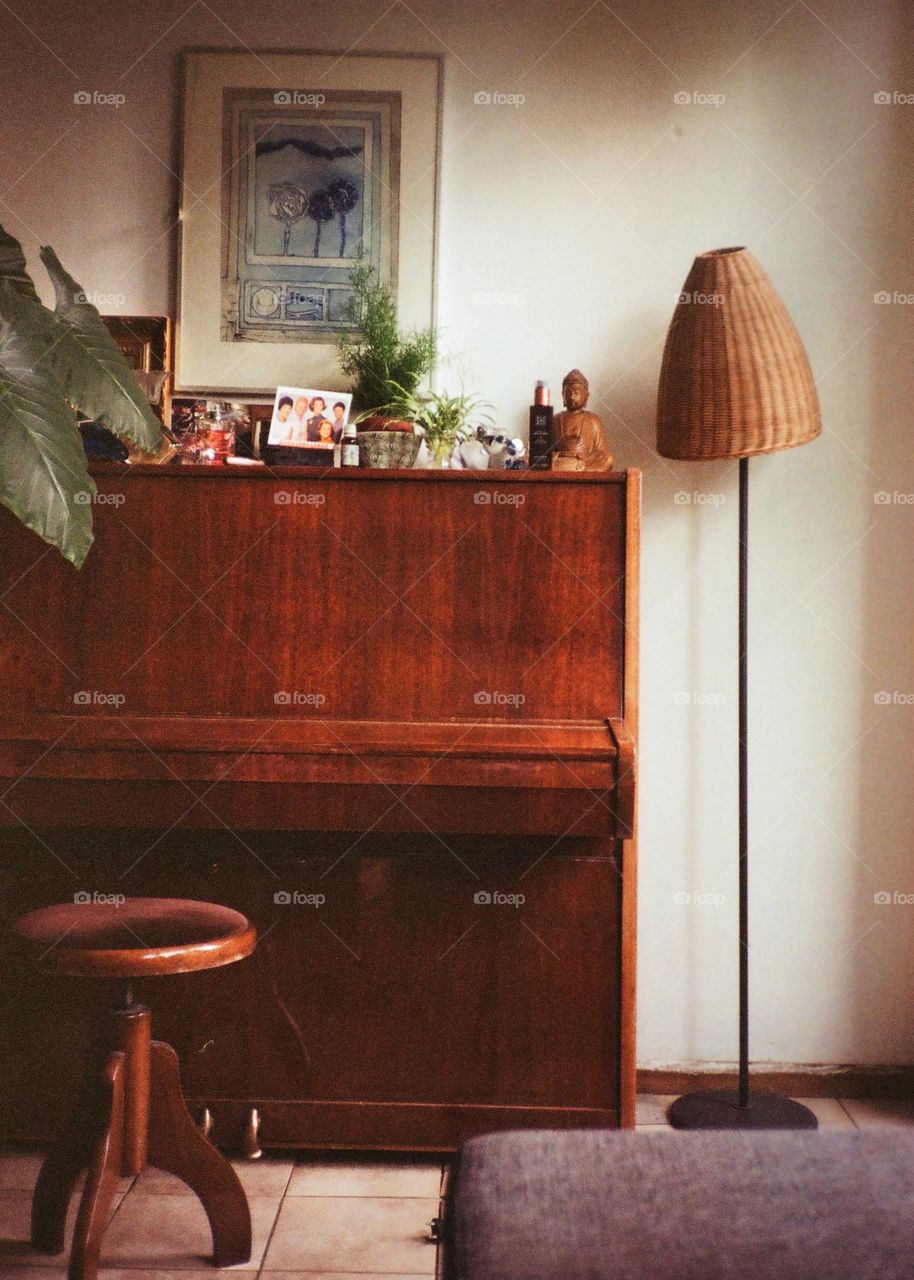 A brown piano and seat with a standing lamp in the corner of a room. Cosy and warm. Vintage feel. Analog photography.