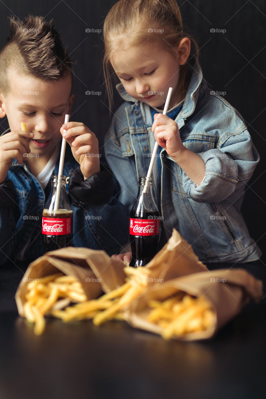 beautiful, stylish, fair-haired children, drinking Coca Cola, eating potatoes, dressed in denim clothes, free photo style. boy and girl have fun, rejoice, beautiful advertisement for coca cola drink