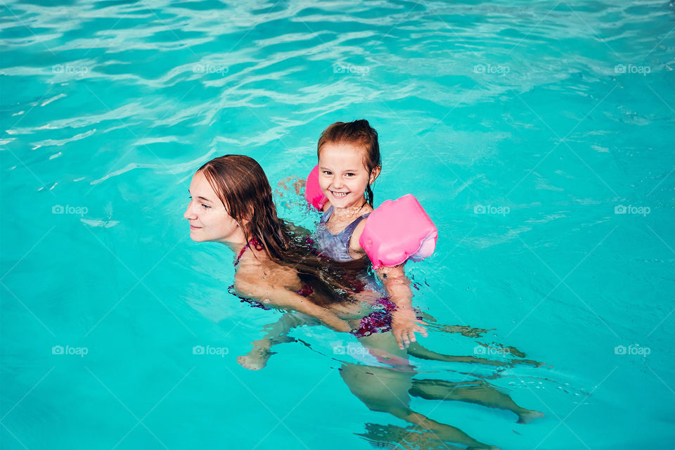 Young woman teaching to swim her younger sister and playing with her in swimming pool. Candid people, real moments, authentic situations