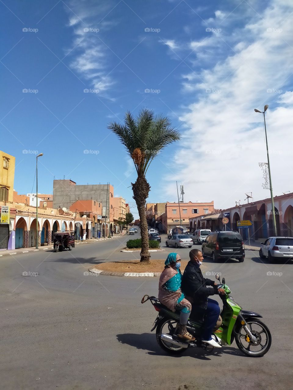 A street through the Souk in Goulimine .we see some arches on the shops ,a man and a woman on a motorbike...a palm tree ...cars...