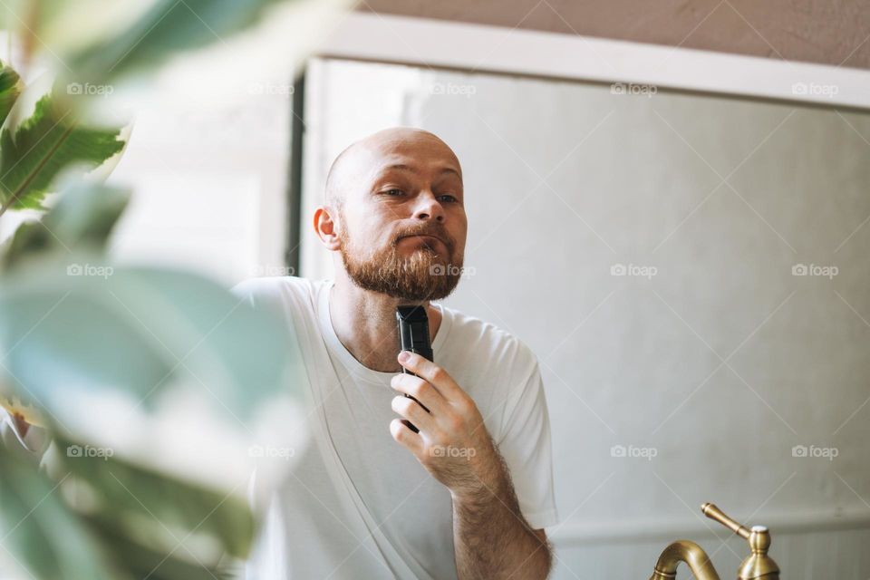 Handsome young bearded man trimming his beard with machine in bathroom at home