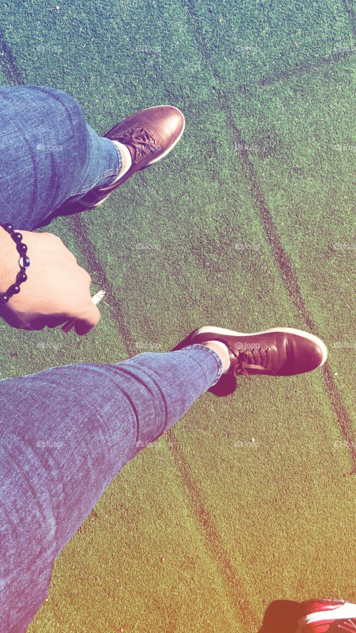 Siting after being tired of walking and smoking a cigarette,black bracelet blue jeans and a brown shoes with small white socks , feet are on the grass 