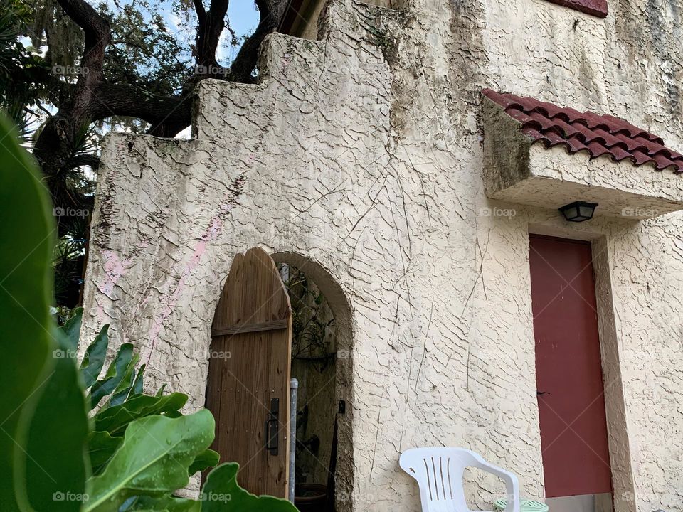 White and red spanish old style architecture residential large house built in the early 1900s. Doorway with antique style wood door in a stairway wall with white chair by a red door seen from tropical plants and live oak trees.