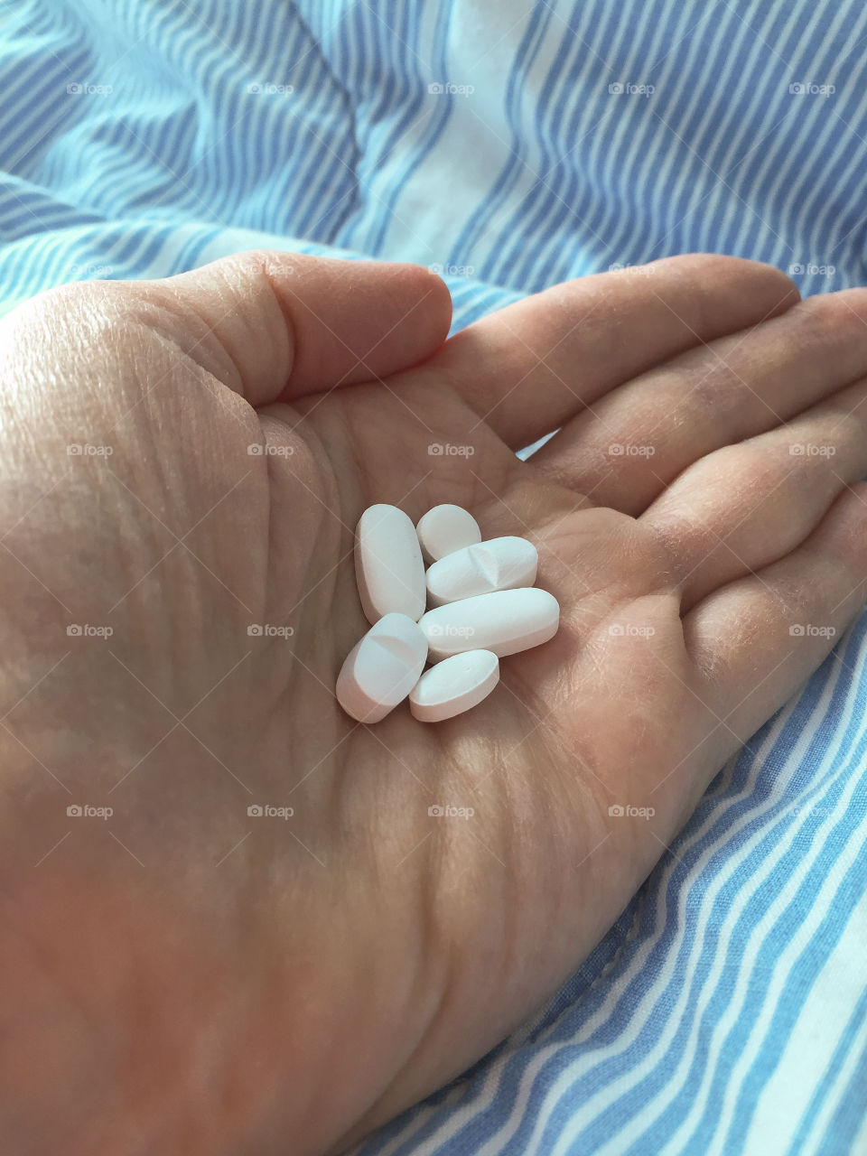 Womens hand holding six white pills in front of a hospital bed with a striped quilt.