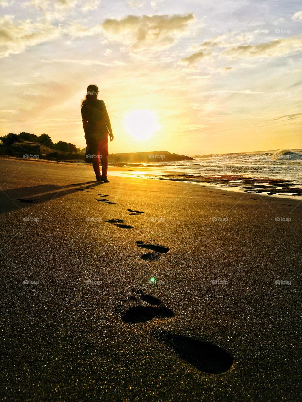 A woman walking in solitude towards the sun on a beach. Her foot prints are the only thing left behind. That too to be washed away by the waves. 