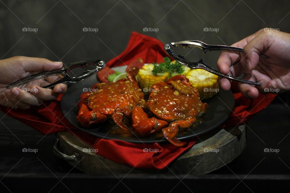 Crab flavored in creamy garlic sauce served with corn, tomatoes, lemon wedges and cherries on a black plate set on a red cloth set in a wooden setting. Asian flavor crab.