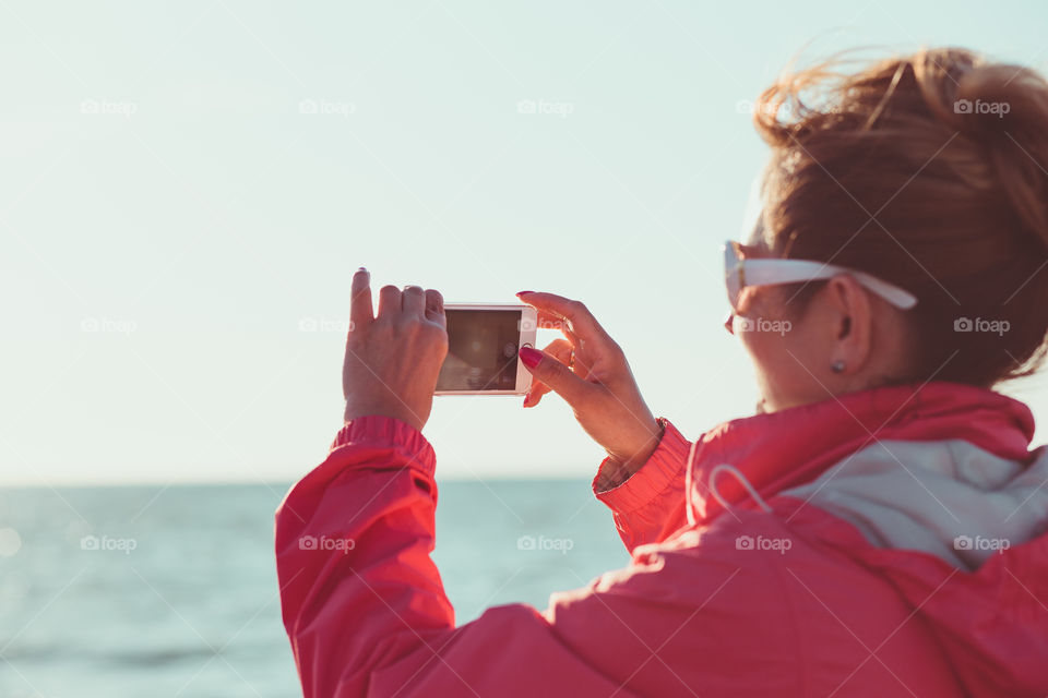 Young woman taking a photos using phone, looking at screen, standing outdoors, she is backlighted by sunlight with plain sky and sea in the background