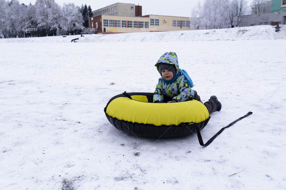 A small, carefree boy walks in the white snow in winter and rides a tubing in the park, near trees in the snow.