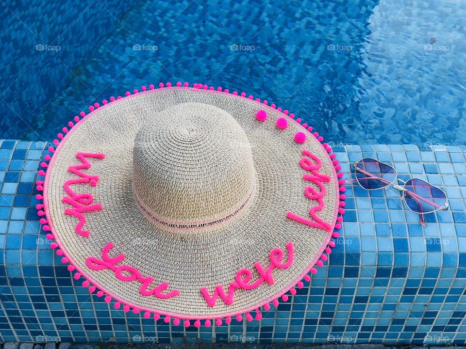 Summer hat with the words “wish you were here” and sunglasses on the edge of a pool with turquoise water