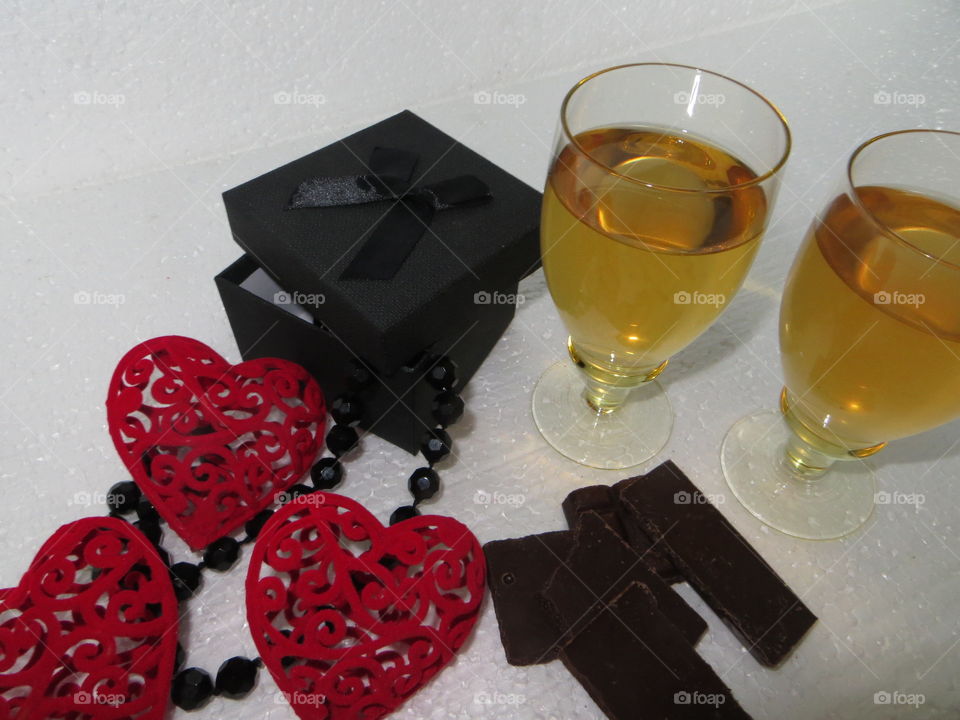 On a white background there are two glasses with white wine, a broken bar of dark chocolate lies next to it, a black gift box with black beads made of agate stone and three bright red tender hearts.