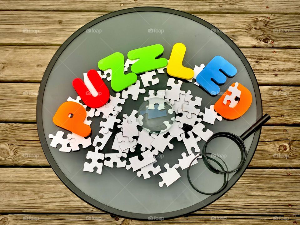 PUZZLED Word Letterings With Puzzle Pieces Centered In The Middle Of Wood Background With Broken Magnifier