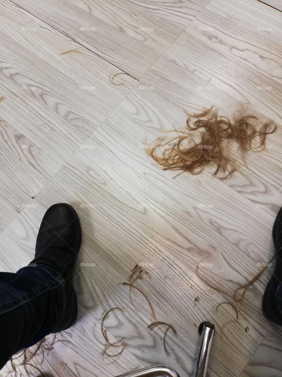 After the hair cut of a blond person the waste is on the floor next to the black shoes and the metal legged chair.