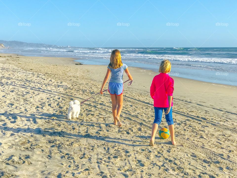 Beach walk! Two girls strolling on the sand, one walking a puppy dog and the other dribbling a soccer ball. 