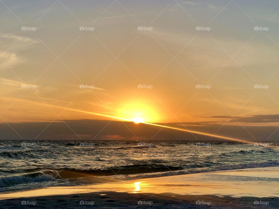 Setting sun over the Gulf of Mexico. The empty beach reflects the golden glow and sunbeam as the sun drops behind a low cloud at the horizon.