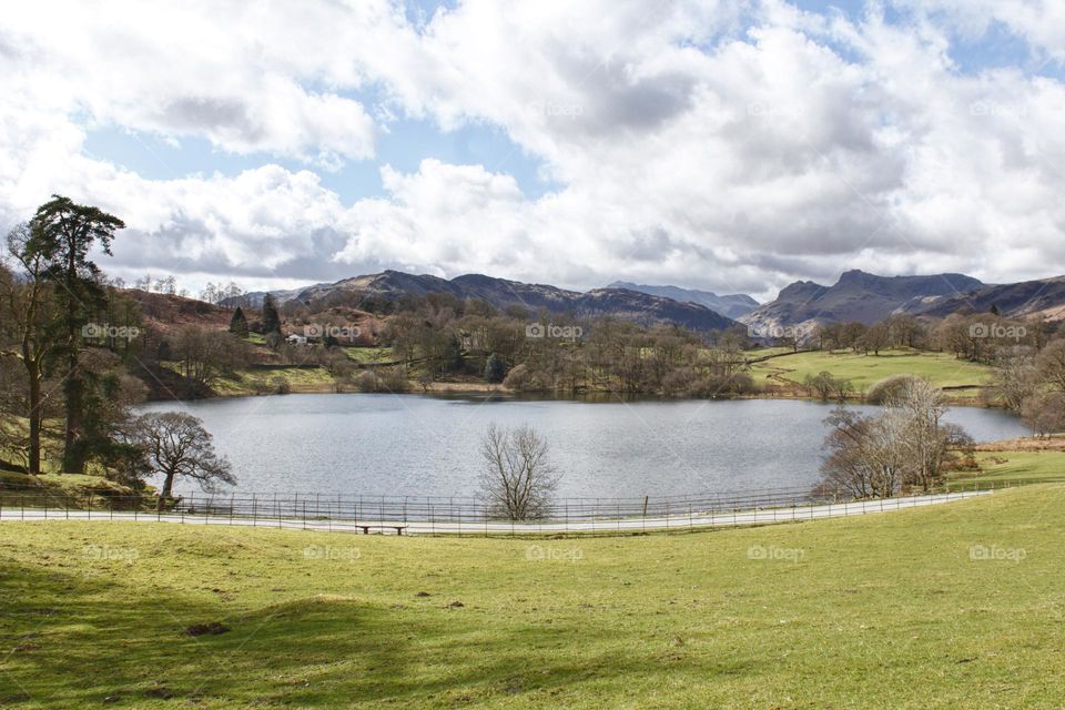 Lake in Lake District in the UK with grass, cloudy sky and mountains in the background.