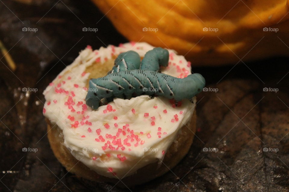 Vanilla cupcake shaped like a donut and adorned with three turquoise blue red-horned hornworms and pink sprinkles. 