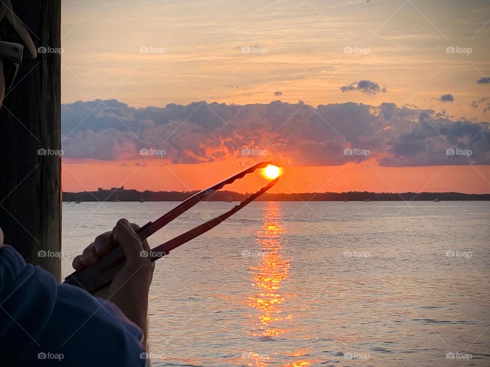 Sun From Sunset Seems To Be Grabbed By The BBQ Tongs  On Top Of An Island In The Horizon At The Seashore On A Tropical Environment.