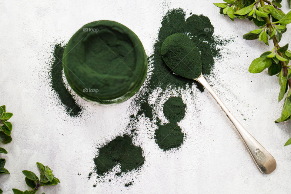 Green powder spirulina powder in a jar with heaped spoon and spills around bottle. Fresh leaves around the green powder
