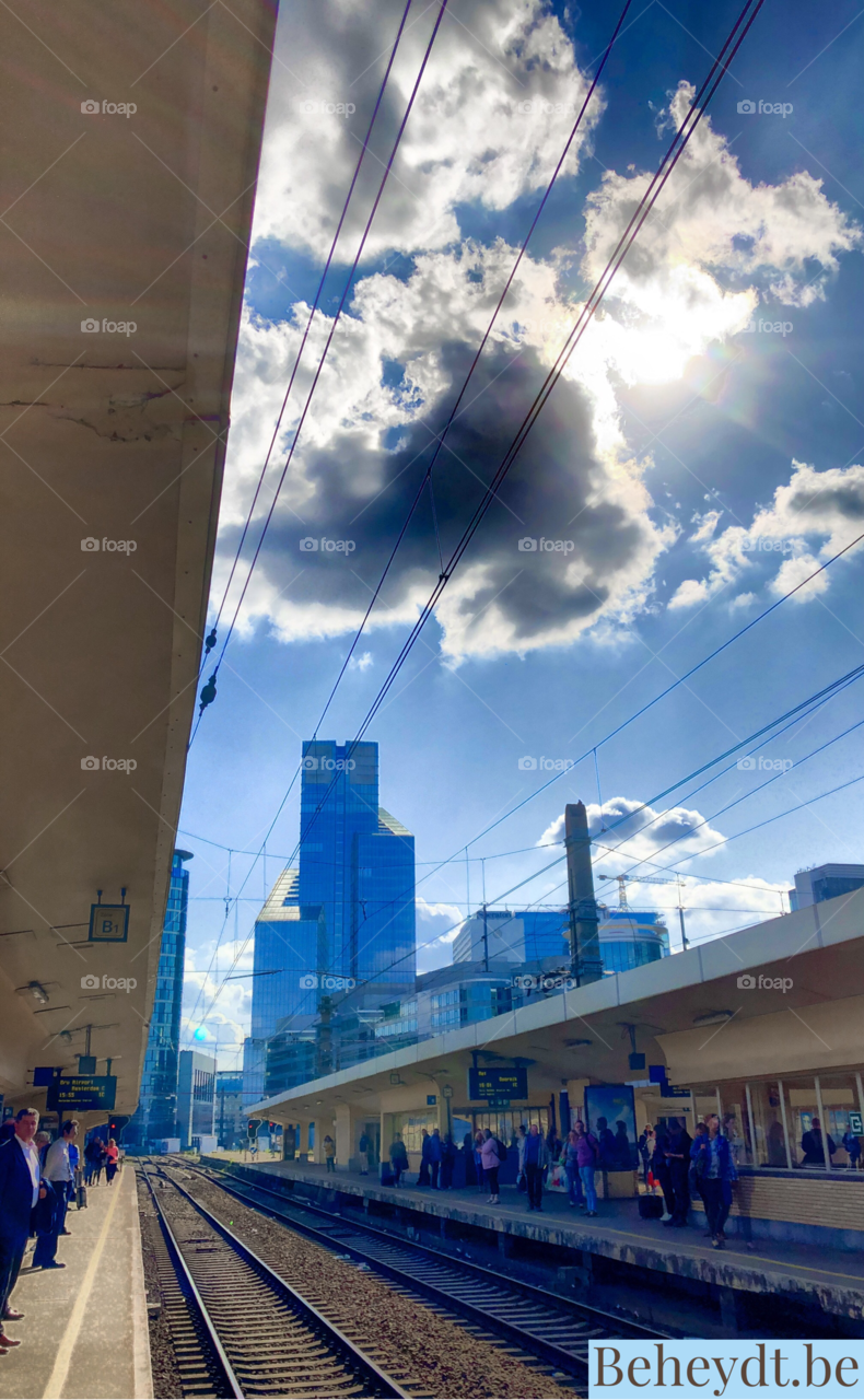 People on the platform of Brussel North railway station waiting for the train under the sunny blue sky with the skyscrapers of Brussels city in the background 