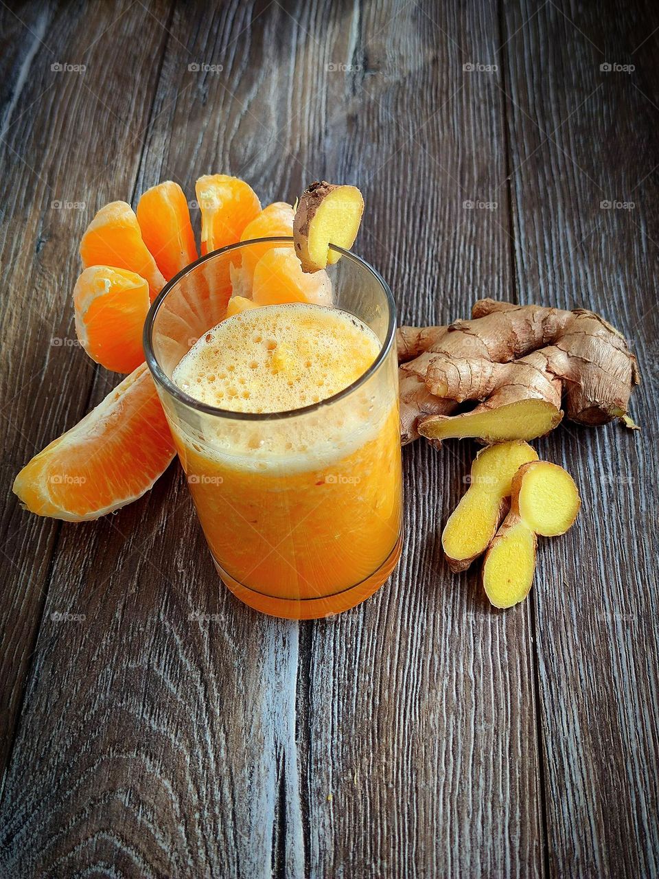 There is a glass with an orange smoothie on a wooden surface. On the glass is a piece of ginger as a decoration. Ingredients lie nearby: peeled orange, ginger root and chopped ginger