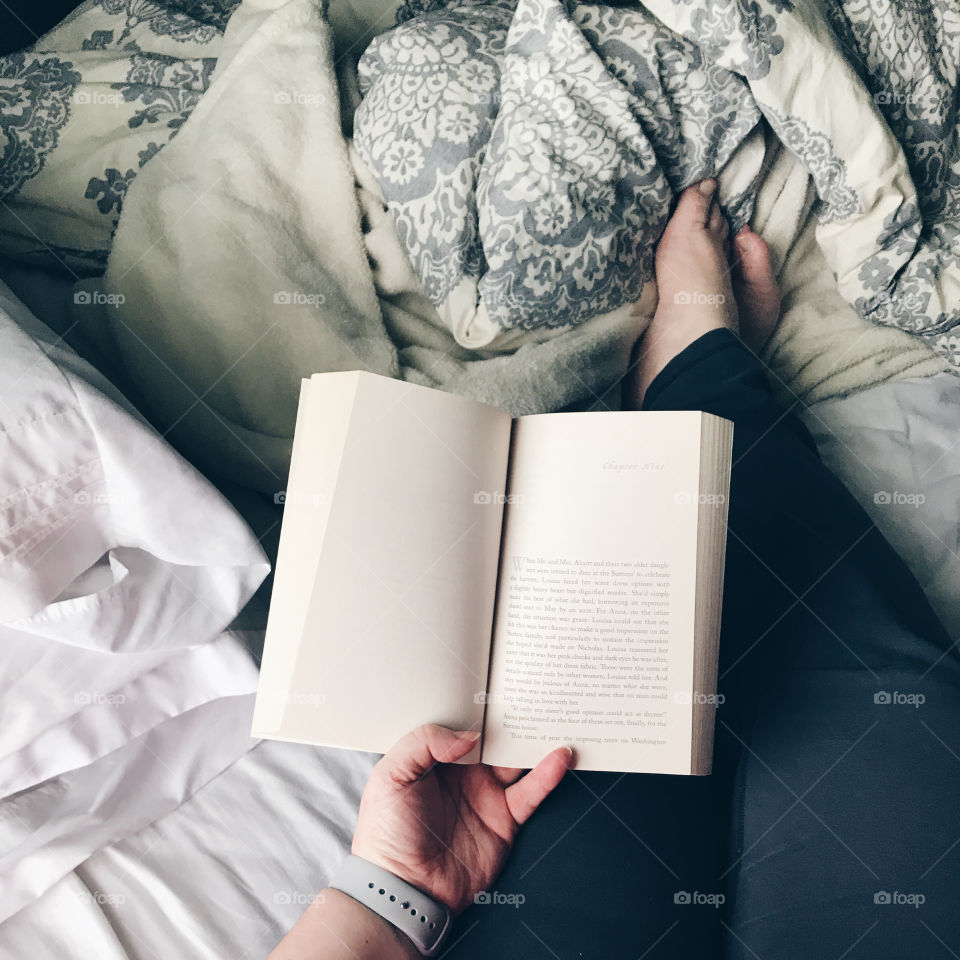 Relaxing scene of a person reading in bed, legs outstretched, bare feet, unmade bed with visible pillows and blankets, hand holding open book
