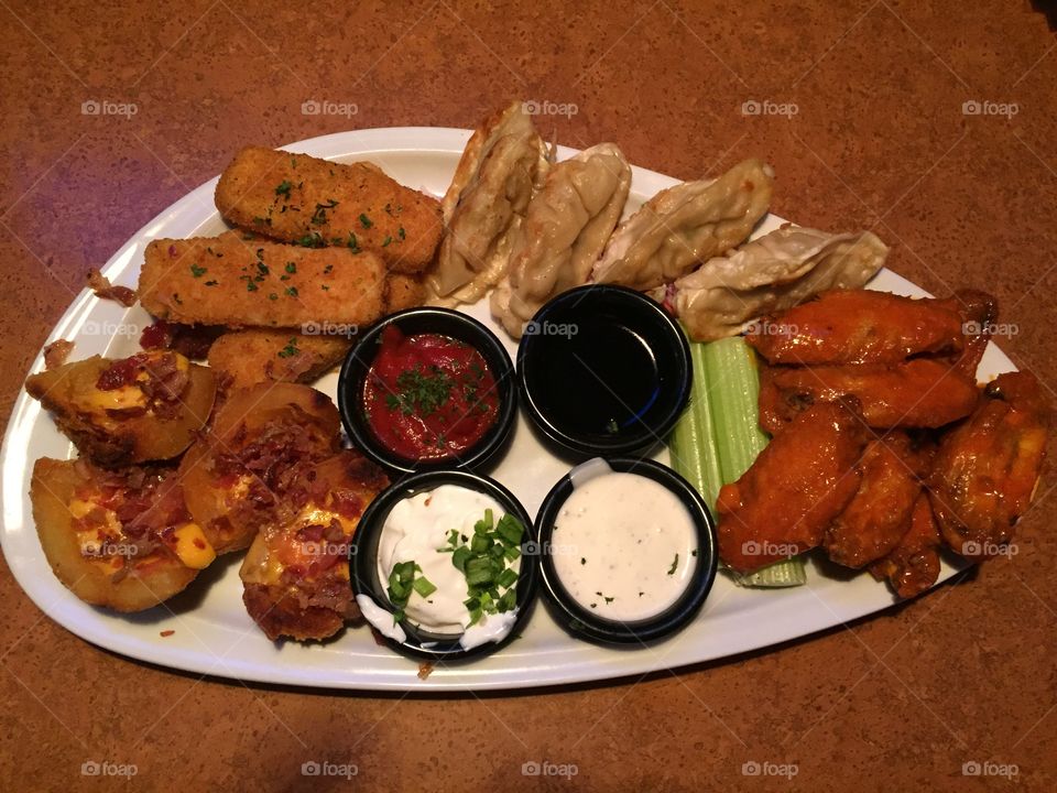 A tasty combination of comfort food consisting of spicy hot wings, potstickers,bacon cheese stuffed potatoes,cheese sticks,,sour cream and chives, a variety of sauces, celery sticks on a platter