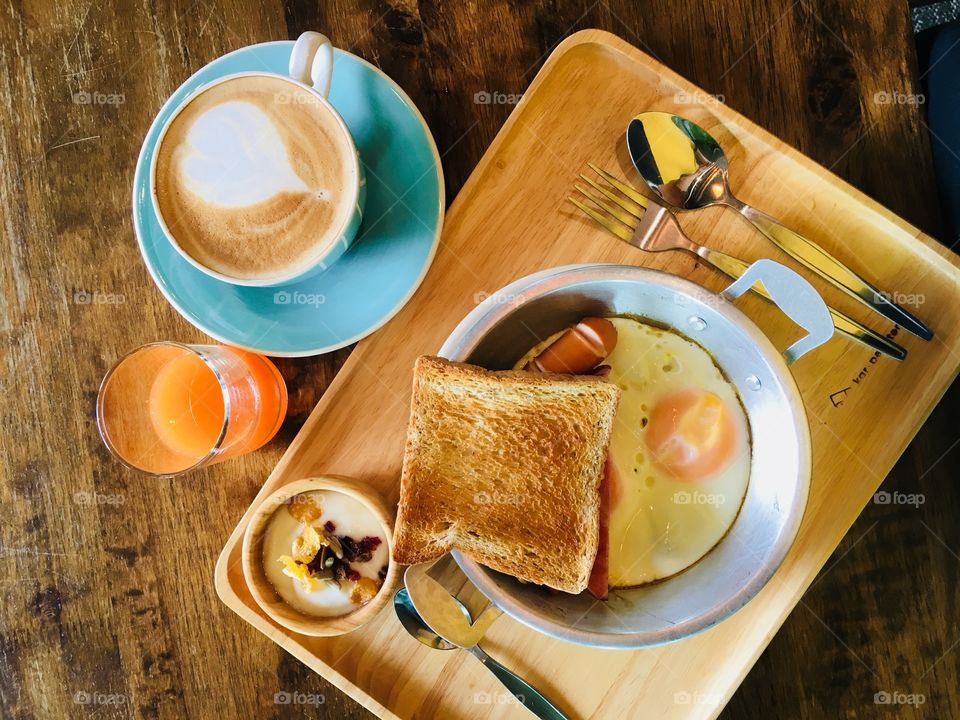 American breakfast fried egg, ham, sausage, bread, yogurt, coffee latte and orange juice serve on wood tray in the morning 